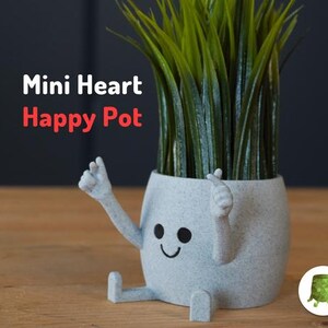 May include: A white ceramic planter shaped like a happy face with arms and legs, holding a small green plant. The planter has the text "Mini Heart Happy Pot" on the background.