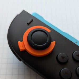 Peut inclure: Gros plan sur une manette de jeu noire et orange. La manette comprend un joystick noir avec un anneau orange et une base noire. La manette comporte également quatre boutons noirs avec des symboles de flèche blancs.