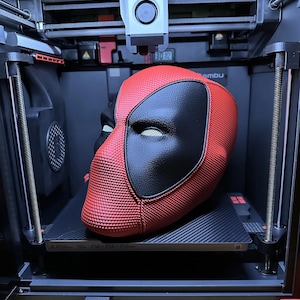 May include: A red and black 3D printed Deadpool mask is being printed on a 3D printer. The printer is a Bambu Lab model. The print bed is 256 x 256 x 256 mm.