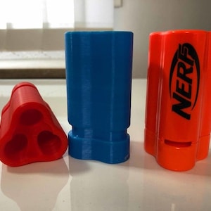 May include: Three plastic toy accessories in red, blue, and orange. The red piece has three holes. The blue piece is a tall cylinder. The orange piece has the word "NERF" printed on it.