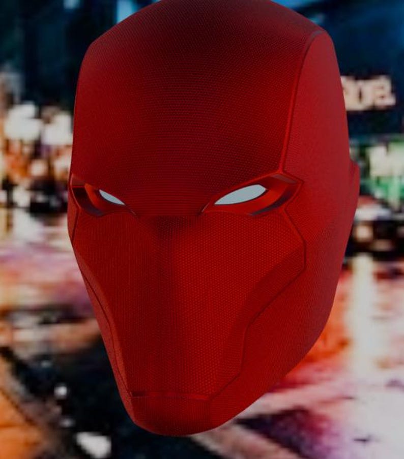 May include: A close-up of a red superhero mask with white eye openings. The mask has a textured surface and a pointed design around the chin and neck. The background is blurred, suggesting an urban setting.