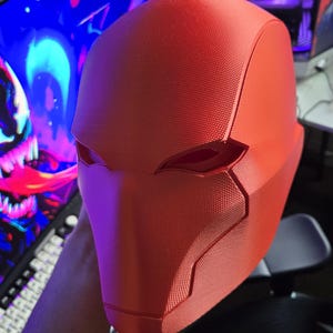 May include: A bright red, textured helmet with angular design elements. The helmet features eye openings and a distinct shape, likely for cosplay or costume use. The overall design suggests a superhero or comic book theme.