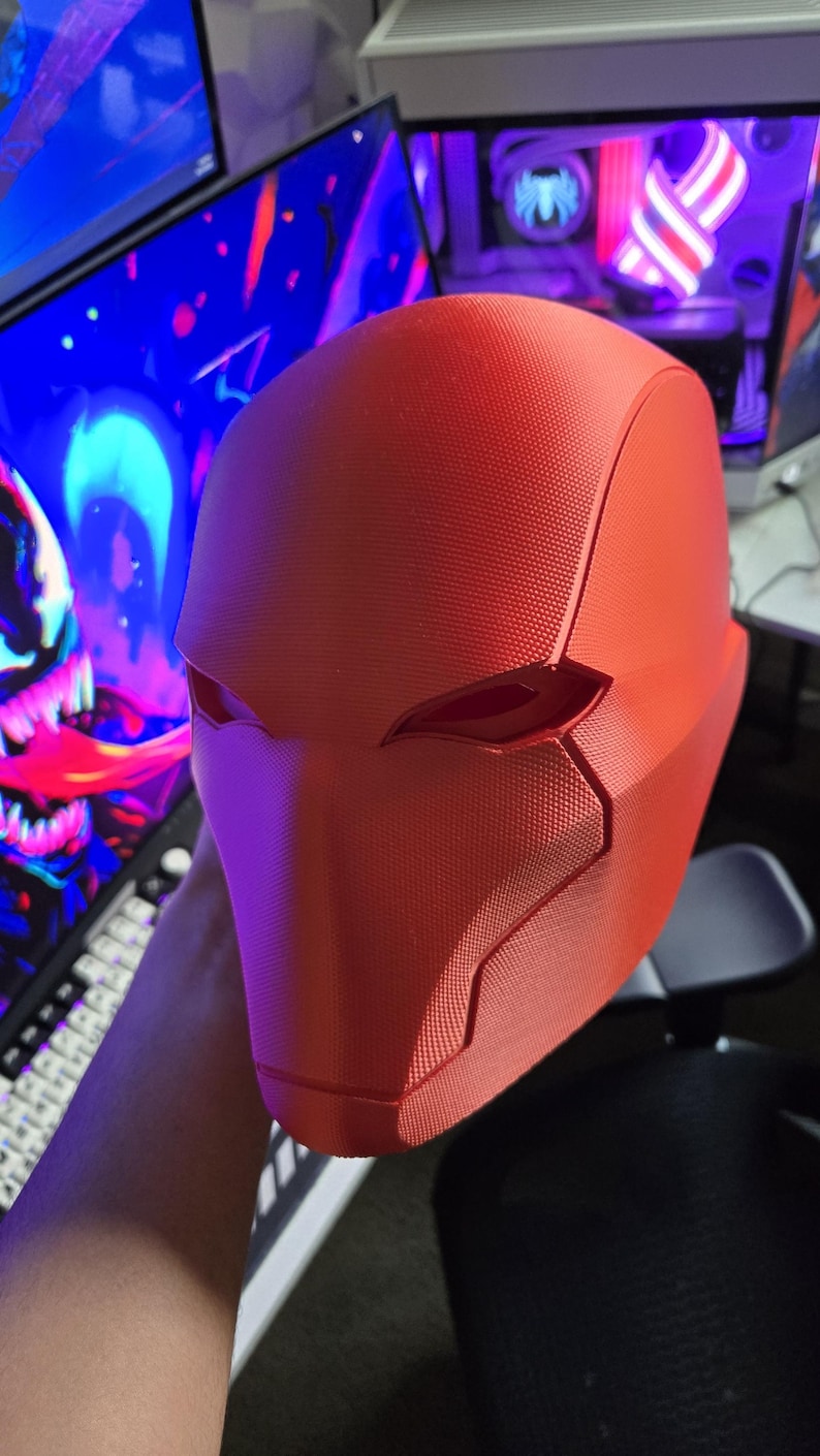 May include: A bright red, textured helmet with angular design elements. The helmet features eye openings and a distinct shape, likely for cosplay or costume use. The overall design suggests a superhero or comic book theme.