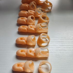 May include: A set of 11 peach-colored plastic finger rings, some with a simple ring shape and others with a more complex design. The rings are arranged in a row on a white surface.