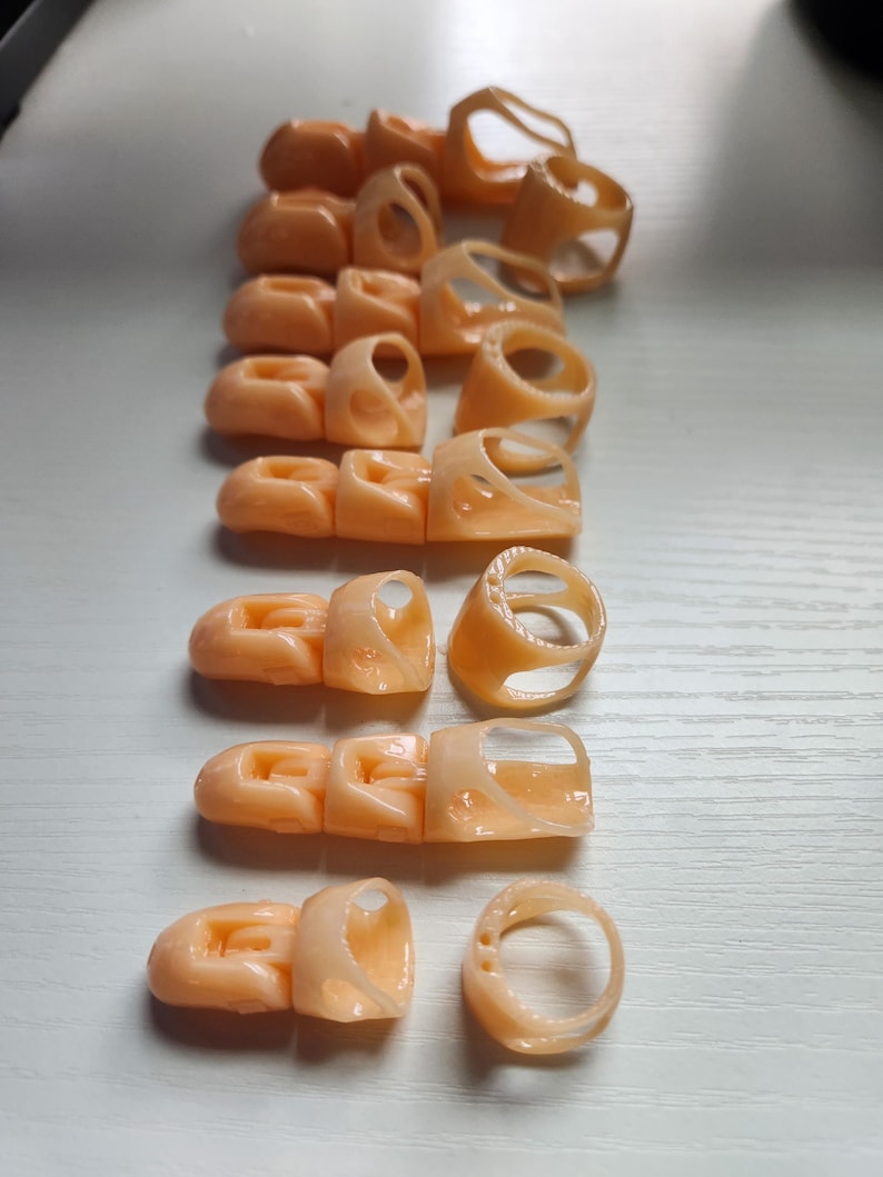 May include: A set of 11 peach-colored plastic finger rings, some with a simple ring shape and others with a more complex design. The rings are arranged in a row on a white surface.