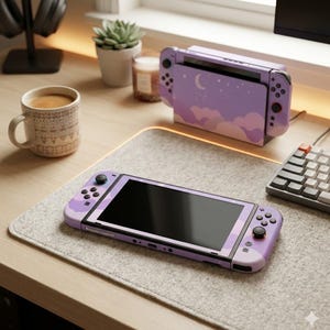 Könnte beinhalten: Eine lavendelfarbene Nintendo Switch-Konsole und Dockingstation mit Wolken- und Monddesign. Die Konsole liegt auf einer grauen Filzmatte, neben einer Kaffeetasse, einer Sukkulente und einer Kerze. Eine Tastatur und Kopfhörer sind ebenfalls zu sehen.