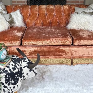May include: A tufted, orange velvet sofa with three seat cushions and rolled arms. The sofa is adorned with decorative pillows and faux fur throws. A cowhide ottoman and a white shag rug are in the foreground.