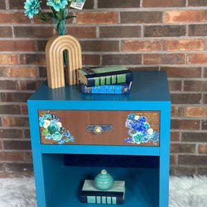 May include: A teal blue nightstand with a drawer featuring floral decals and a brown wood panel. The nightstand has a lower shelf and is decorated with books, a vase, and a decorative wooden arch. The drawer has a small, round knob.