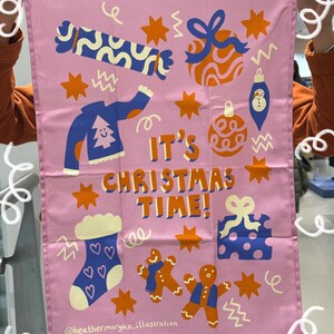 Puede incluir: Paño de cocina rosa con ilustraciones navideñas, incluyendo un suéter, cajas de regalo, adornos y hombrecitos de jengibre. El texto "IT'S CHRISTMAS TIME!" se muestra de forma destacada. También es visible el nombre del artista.