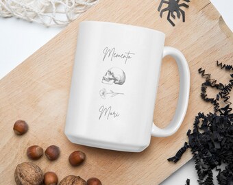 May include: White ceramic mug with a black and white illustration of a skull and a rose. The text "Memento Mori" is printed on the mug.