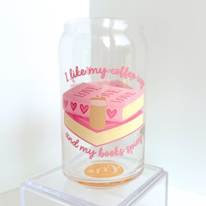 May include: A clear glass tumbler featuring a pink graphic of two stacked books with hearts and a coffee cup. The text reads I like my coffee icy and my books spicy in pink script lettering. The design is printed on the front of the cylindrical glass.