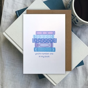 May include: A white greeting card featuring a stack of illustrated books in blue and purple hues. The card displays the text "you're number one in my book." It rests on a pile of books and a brown envelope.