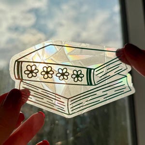 Puede incluir: Una pegatina transparente con una pila de dos libros con un diseño floral. Los libros tienen un acabado holográfico iridiscente, y los lomos están decorados con tres flores. La pegatina se sostiene contra una ventana.