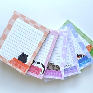 May include: A collection of lined notepads with cat illustrations. Each notepad features a different cat design, including a black cat, a calico cat, and a tabby cat. The notepads have patterned borders in shades of pink, purple, and green.