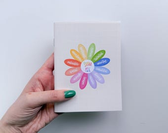 May include: A white notebook with a faint grid pattern featuring a colorful flower graphic on the cover. The center of the flower reads "YOU ARE" surrounded by petals labeled with words including "valuable," "special," "enough," "brave," "capable," "strong," "beautiful," "worthy," "talented," "lovable," "brilliant," and "wonderful."