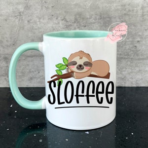 May include: White ceramic mug with a light green handle and interior. The mug features a cartoon sloth resting on a branch with green leaves, and the word "SLOFFEE" in black lettering.
