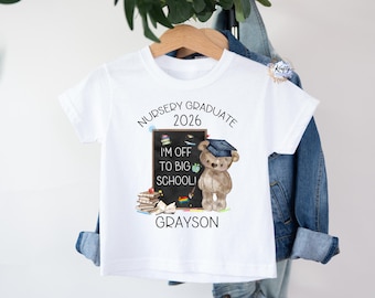 May include: White t-shirt with a graduation-themed graphic. The design includes a teddy bear wearing a graduation cap, a chalkboard with text, and the name "Grayson." The text reads "Nursery Graduate 2026" and "I'm off to big school!"