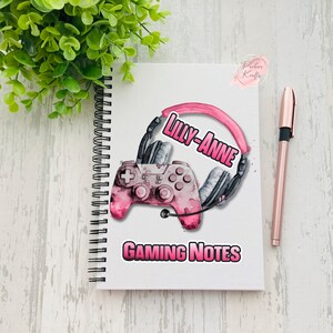 May include: White spiral-bound notebook with a pink and grey watercolour design featuring a gaming controller and headset. The text "Lilly-Anne" and "Gaming Notes" are displayed in pink. A rose gold pen rests beside the notebook.