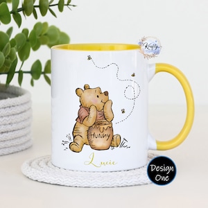 May include: White ceramic mug with a yellow rim and handle. The mug features a cartoon illustration of Winnie the Pooh holding a honey pot. The text "Hunny" is on the honey pot. The text "Lucie" is at the bottom of the mug.