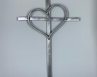 May include: A silver metal cross with a heart-shaped cutout in the center. The cross is made of thick metal and has a simple, elegant design.