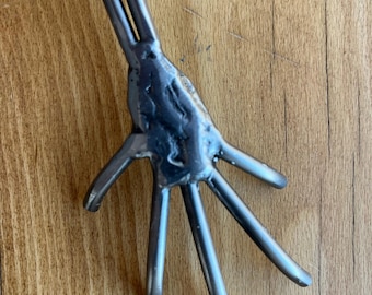 May include: A handcrafted metal garden tool with a hand-shaped design. The tool features five finger-like tines extending from a textured palm, and two long, parallel handles. The metal has a silver finish.