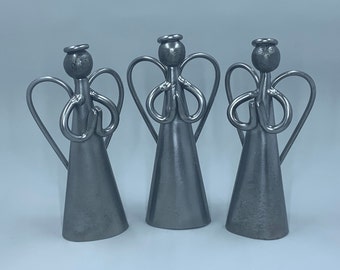 May include: Three silver metal angel figurines with wings and hands clasped in prayer. The angels are standing on a white surface.