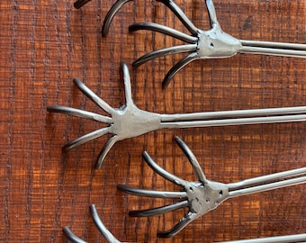 May include: A set of five metal roasting sticks, each featuring a hand-shaped end with five long, curved fingers. The metal is dark grey with visible weld marks at the base of the fingers, and each hand is attached to a long, straight metal rod.