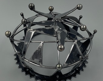May include: A metal sculpture in the shape of a crown with a circular base and a star-shaped top. The crown is made of recycled metal and has a rustic, industrial look.