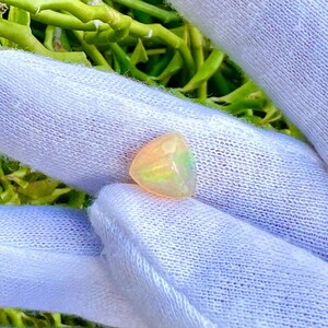 May include: A white gloved hand holding a small, triangular, iridescent opal gemstone. The opal has a light yellow, green, and blue color.