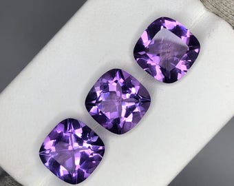 Könnte beinhalten: Drei quadratisch geschliffene, facettierte Amethyst-Edelsteine. Die Edelsteine haben eine tiefviolette Farbe und werden auf einer weißen Oberfläche präsentiert. Die Facetten fangen das Licht ein und erzeugen einen funkelnden Effekt. Die Edelsteine sind in einer Reihe angeordnet.