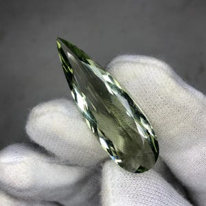 May include: A teardrop-shaped green gemstone with a faceted cut. The gemstone is held in a white gloved hand.