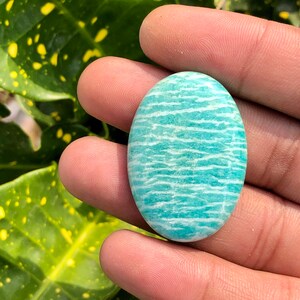 Puede incluir: Gema ovalada de amazonita, que muestra un color azul verdoso claro con bandas horizontales blancas. La piedra pulida se sostiene en una mano, resaltando su superficie lisa y sus patrones naturales. La gema es una piedra natural.