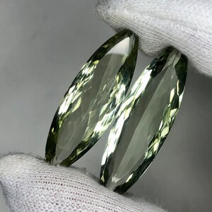 May include: Two light green marquise cut gemstones, set on a white fabric background.
