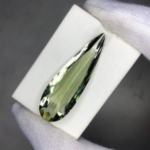 May include: A pale green, elongated gemstone with multiple facets. The gem is displayed on a white surface, highlighting its polished finish and transparency. The stone's shape is similar to a teardrop or a narrow leaf.