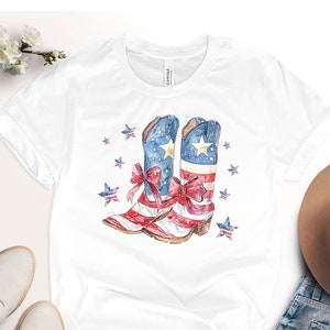 May include: White t-shirt featuring a watercolour design of red, white and blue cowboy boots with a bow on each boot. The boots are decorated with stars and are reminiscent of the American flag.