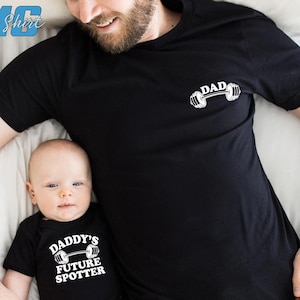 May include: A black t-shirt with the text "DAD" and a barbell graphic. A black bodysuit with the text "Daddy's Future Spotter" and a barbell graphic. A father and child are wearing the shirts.