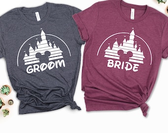 May include: Two t-shirts with a white silhouette of Cinderella's castle with Mickey Mouse ears in the center. The shirt on the left is dark gray with the word "Groom" below the castle. The shirt on the right is burgundy with the word "Bride" below the castle.