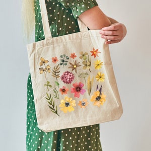 May include: A beige tote bag with a floral design featuring a variety of colorful flowers and leaves. The bag has two long straps and is being held by a person wearing a green polka dot dress.
