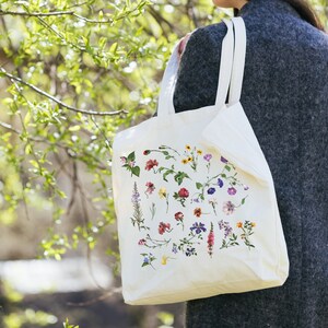 Peut inclure: Un sac fourre-tout blanc avec un imprim&eacute; floral repr&eacute;sentant diff&eacute;rentes fleurs dans des tons de rose, violet, jaune et vert. Le sac a deux longues poign&eacute;es et est tenu par une personne.