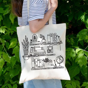May include: A white tote bag with black line drawings of a bookshelf with plants, books, and a globe. The bookshelf has two shelves and is decorated with string lights.