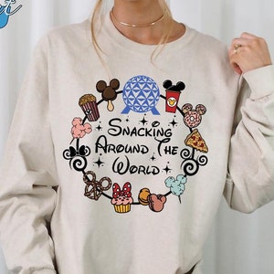 May include: A beige long-sleeve shirt with a graphic of a circle of Disney snacks and characters, including Mickey Mouse, Minnie Mouse, and the Spaceship Earth ride. The text "Snacking Around the World" is inside the circle.