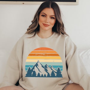 May include: A beige sweatshirt featuring a vintage-style graphic of mountains and a sunset. The graphic is in a blue, green, yellow, orange, and red colour scheme.