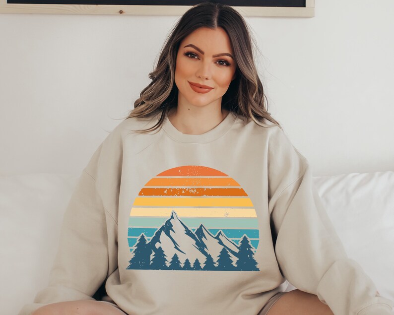 May include: A beige sweatshirt featuring a vintage-style graphic of mountains and a sunset. The graphic is in a blue, green, yellow, orange, and red colour scheme.