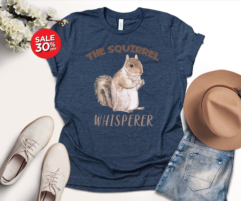 May include: A navy blue t-shirt with the text "The Squirrel Whisperer" in brown lettering. A watercolor illustration of a squirrel is in the center of the shirt.