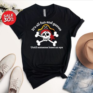 May include: Black t-shirt with a white skull and crossbones graphic. The skull is wearing a red bandana and a pirate hat. The text "It's all fun and games Until someone loses an eye" is printed below the graphic.