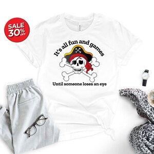 May include: White t-shirt with a black and white skull and crossbones graphic. The skull is wearing a red bandana and a black pirate hat. The text "It's all fun and games Until someone loses an eye" is printed below the graphic.