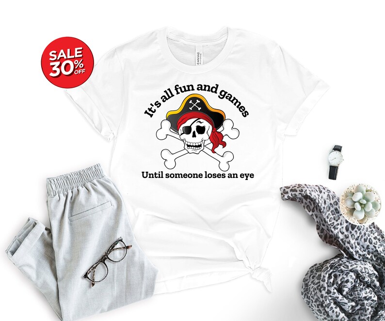 May include: White t-shirt with a black and white skull and crossbones graphic. The skull is wearing a red bandana and a black pirate hat. The text "It's all fun and games Until someone loses an eye" is printed below the graphic.