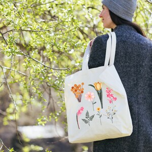 May include: A white canvas tote bag with a floral design. The bag features a variety of flowers and plants in shades of pink, orange, and yellow. The bag is being held by a person wearing a gray beanie and a gray sweater.