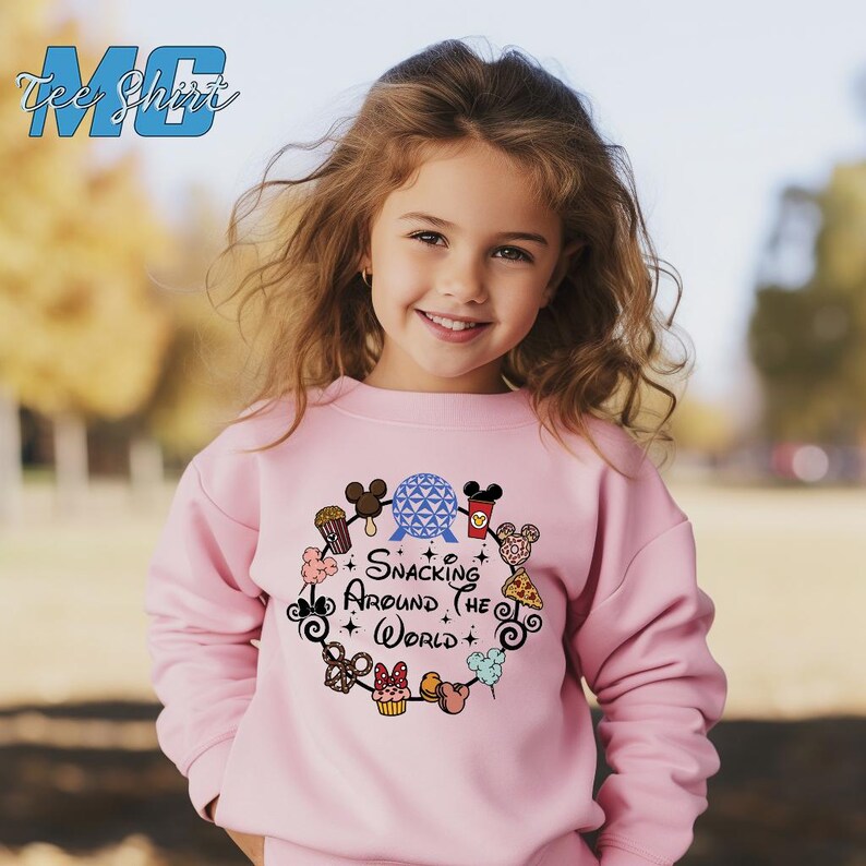 May include: A pink sweatshirt with a graphic of a blue and white sphere surrounded by various Disney characters and snacks. The text "Snacking Around the World" is printed on the sweatshirt.