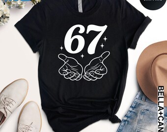 May include: Black t-shirt with the number 67 in white, accompanied by a hand gesture graphic. The shirt is made of a soft material and is suitable for casual wear. The design includes small star accents.